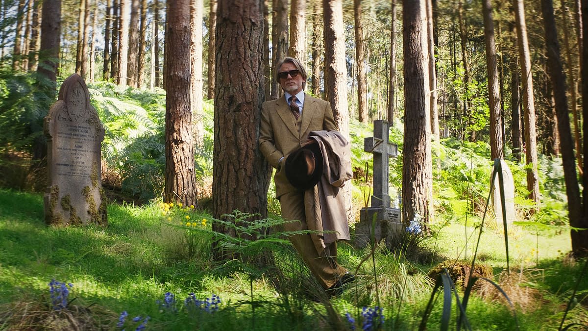 A still from 'Wake Up Dead Man: A Knives Out Mystery,' featuring a man in a tan suit and sunglasses leaning against a tree in a graveyard, surrounded by gravestones and lush greenery.