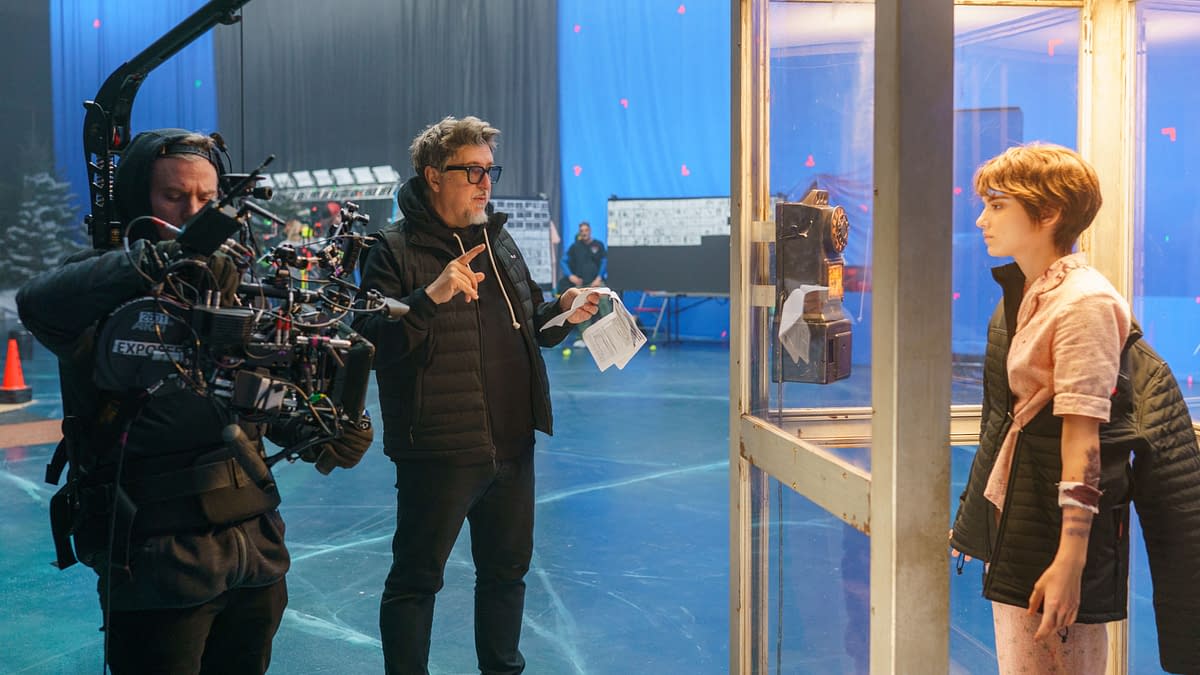 Behind-the-scenes image from 'Black Phone 2' featuring a director guiding the filming process with a camera, while a young actress stands in a glass phone booth next to an old-fashioned payphone.