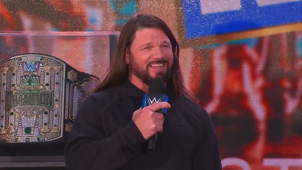 AJ Styles, wearing a black hoodie, smiles while speaking into a microphone at the Crown Jewel Perth Kickoff Show, with a championship belt displayed behind him.