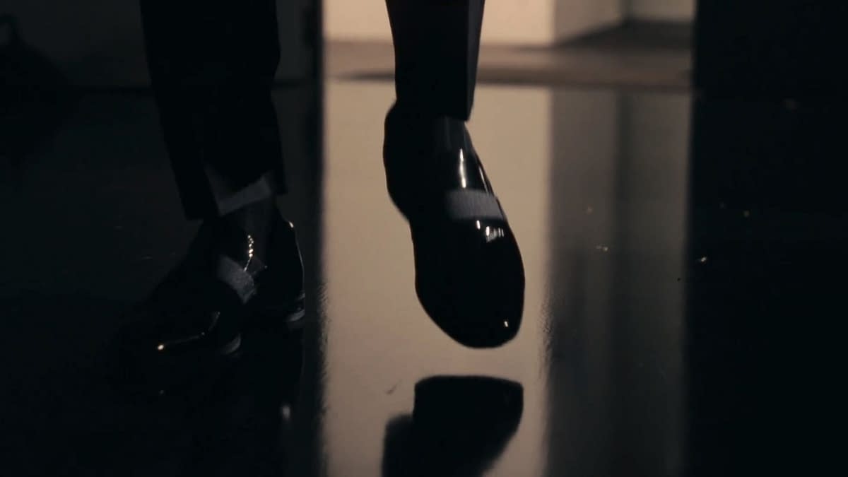 A close-up of a foot wearing a shiny black shoe, poised to take a step on a reflective surface. The shoe features a strap, emphasizing a sleek design.