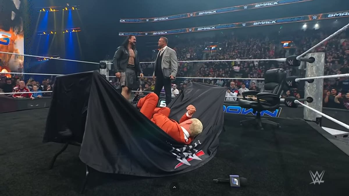 Cody Rhodes lies on the floor, partially covered by a black cloth, after a table break during a WWE SmackDown contract signing with Drew McIntyre. A referee and other officials stand nearby, surrounded by a cheering crowd.