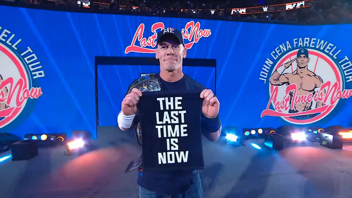 John Cena stands in the center of the wrestling ring at Madison Square Garden, holding a black towel with the words "THE LAST TIME IS NOW" printed in large white letters. He wears a championship belt and a cap, with bright stage lights illuminating the scene.