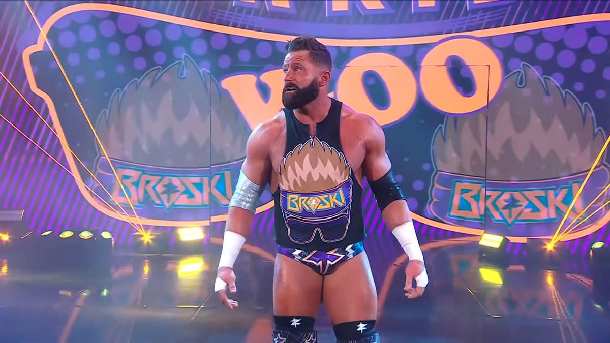 A muscular wrestler stands in the spotlight on a colorful stage, wearing a tank top with a cartoonish design featuring sunglasses and the word 'BROSKI.' The backdrop has bright purple and yellow patterns with the words 'Woo Woo Woo.'