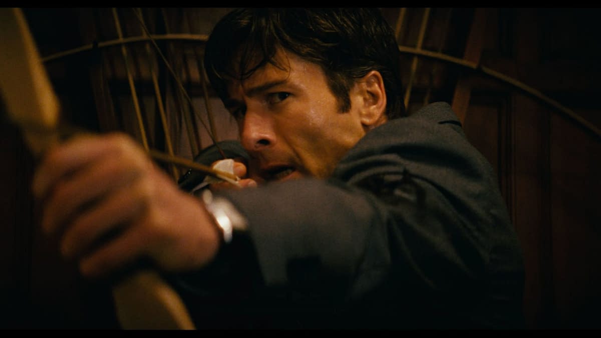 A man in a suit aims an arrow from a bow, displaying intense focus and determination. The background features wooden paneling, adding to the dramatic atmosphere.