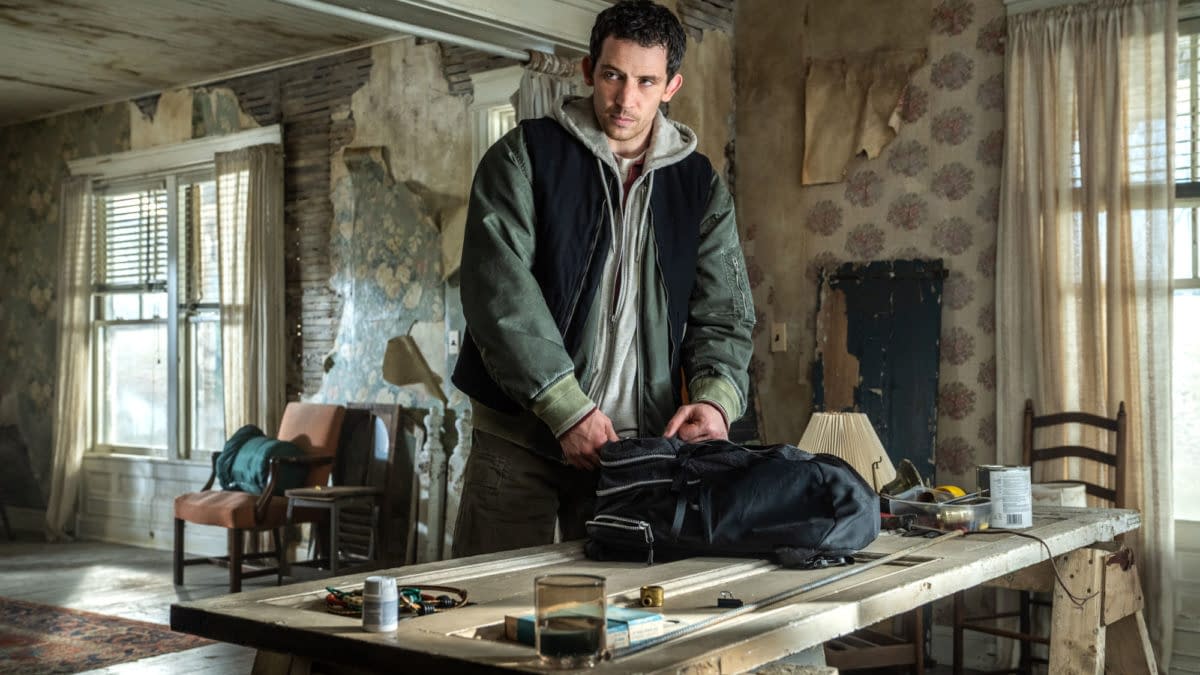 A scene from the film 'Disclosure Day,' featuring a man, Josh O'Connor, packing a black bag on a wooden table in an aged, unkempt room with floral wallpaper and a mix of furniture in the background.