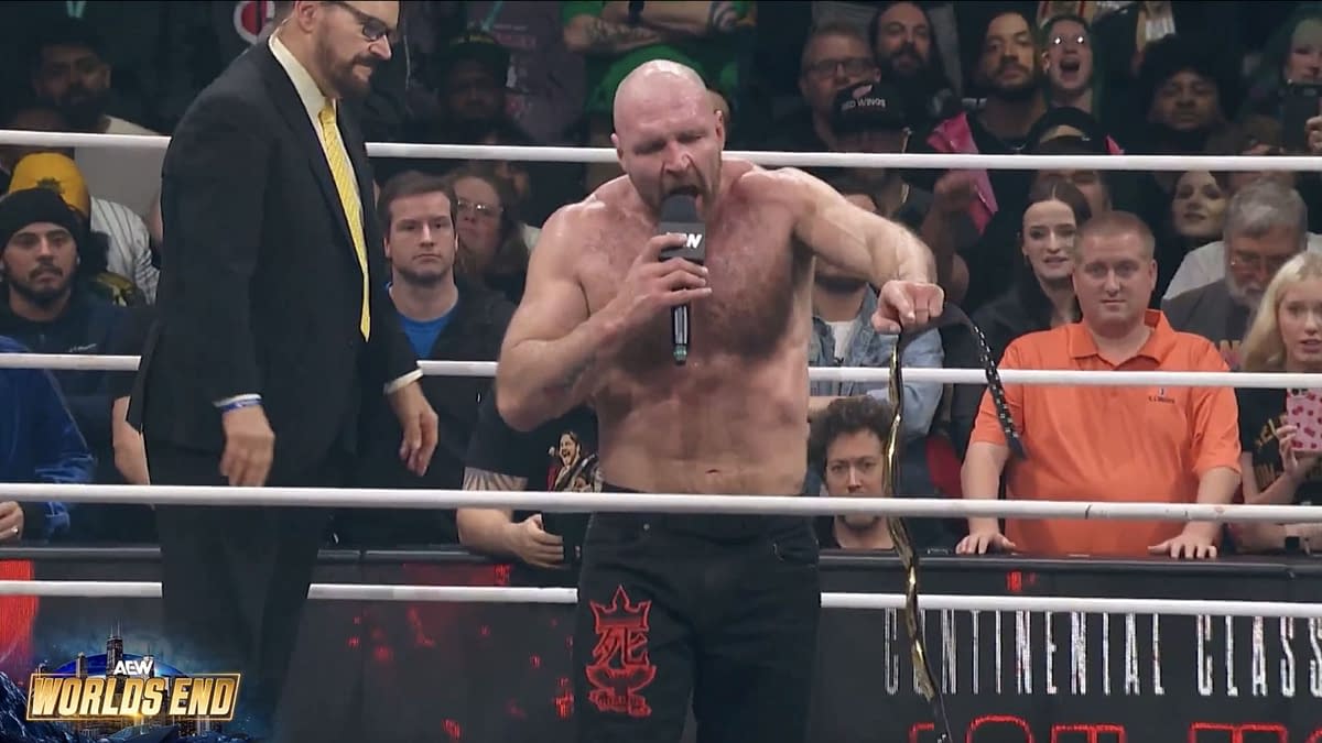 Jon Moxley delivers a passionate promo in the wrestling ring at AEW World's End, holding a championship belt and addressing fans and wrestlers. A commentator stands nearby while the enthusiastic audience is visible in the background.