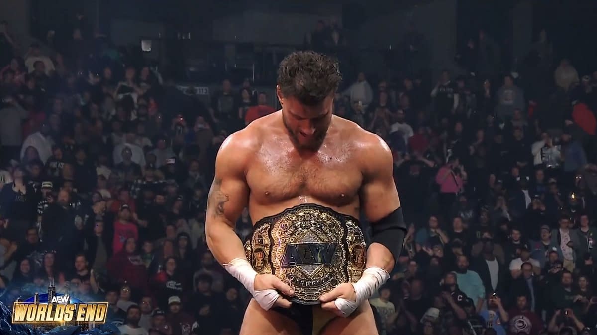 A wrestler stands in the ring, holding the AEW World Championship belt and looking down at it, surrounded by an excited crowd of fans. The atmosphere is electric, reflecting the significance of the moment.
