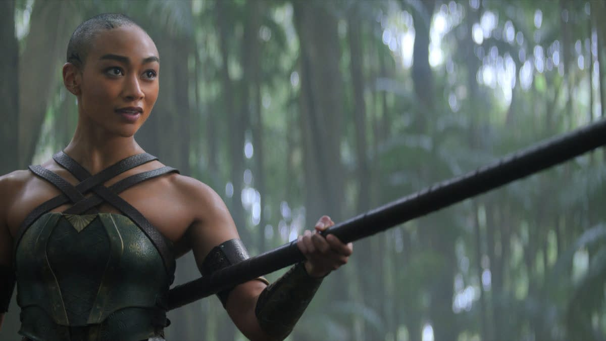 A female character, dressed in a green and gold warrior outfit, stands confidently in a lush forest environment, holding a long spear. The sunlight filters through the trees, creating a mystical atmosphere.