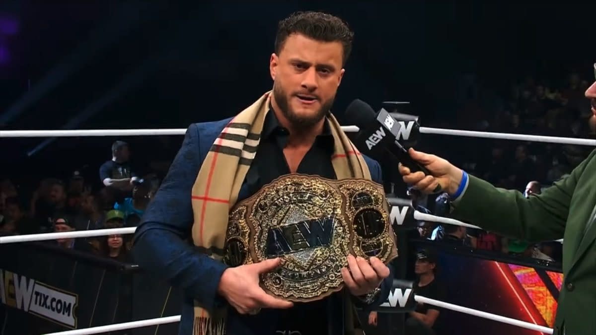 A wrestler holds the AEW World Championship belt while being interviewed by another individual, dressed in a blazer and a plaid scarf. The wrestling ring is visible in the background with an audience.