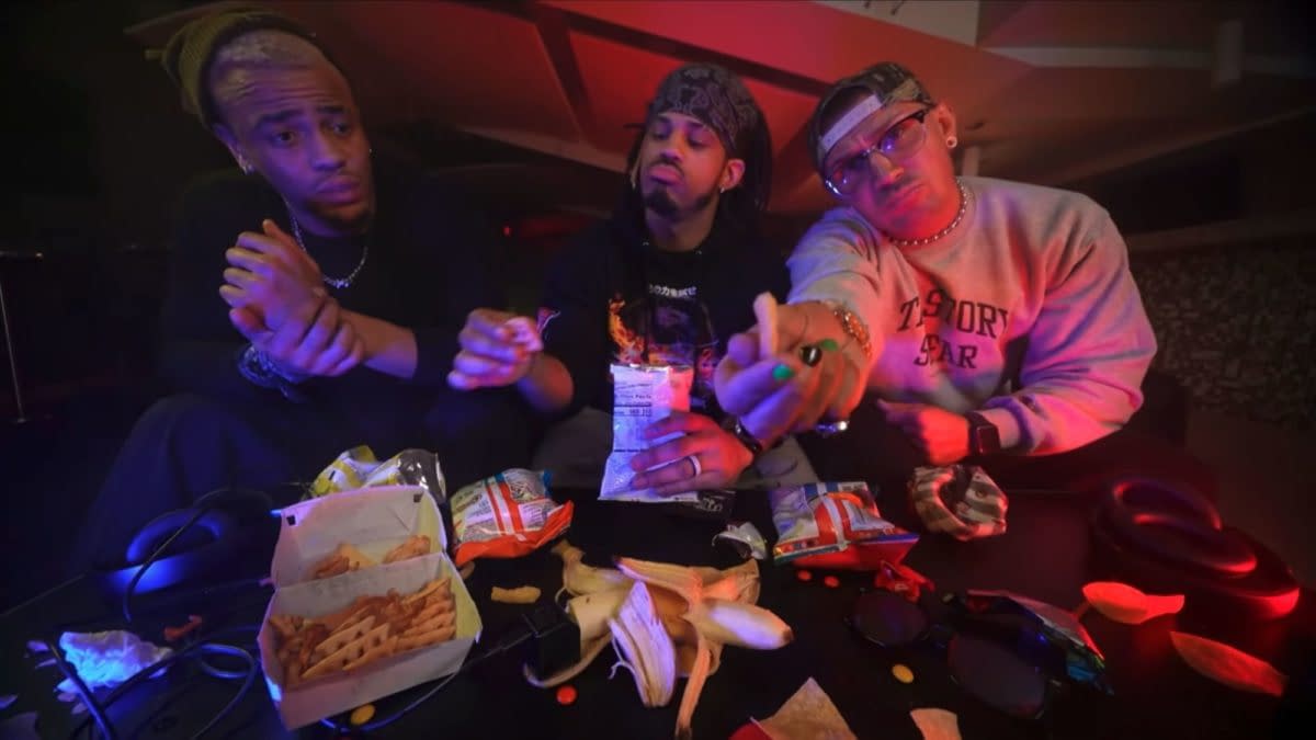 Three members of The Rascalz relax backstage on AEW Collision, seated around a table filled with snacks, including fries and various chips. The dimly lit setting features colorful ambient lighting, highlighting their expressions as they interact with each other.