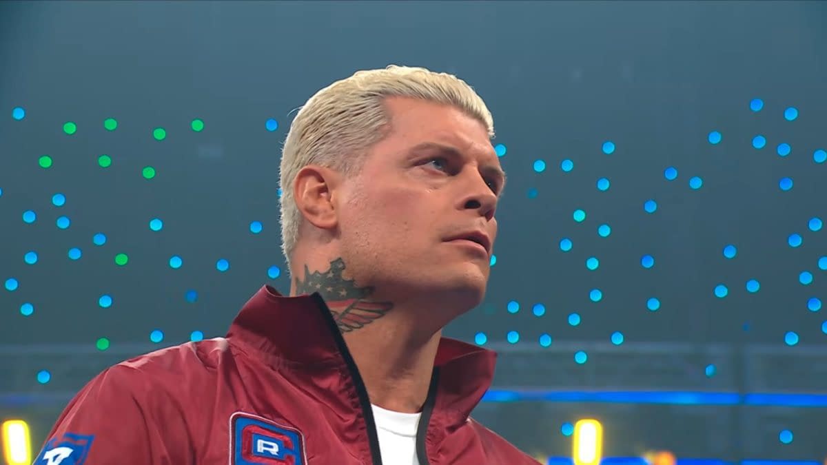 Cody Rhodes appears on WWE SmackDown, showcasing a serious expression and styled hair, wearing a maroon jacket with a logo. The background features colorful lights and a wrestling ring environment.