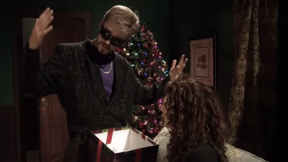 A man in sunglasses and a patterned black jacket gestures dramatically while holding an open gift box, with a Christmas tree lit up in the background. An intrigued woman with curly hair watches.