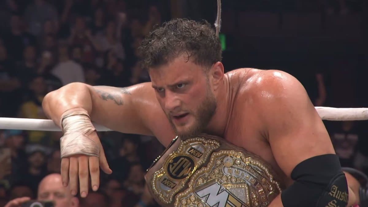 A wrestler, wearing a championship belt and bandaged hand, looks intensely at the audience from the ring at AEW Grand Slam Australia. His sweat glistens as the crowd cheers behind him.