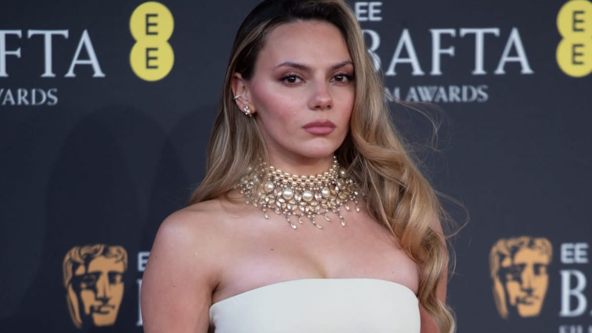 A woman with long, wavy hair wearing a glamorous pearl necklace and a strapless white gown poses confidently at the BAFTA Film Awards backdrop.
