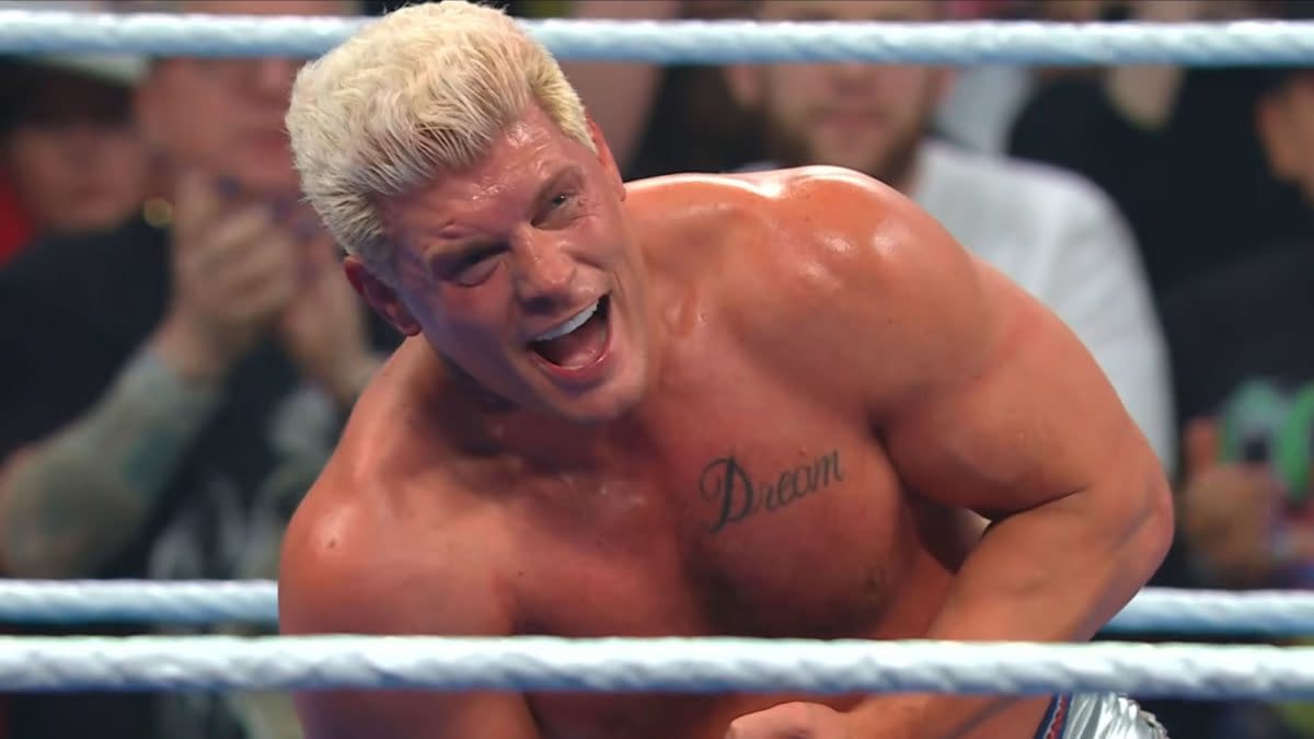 Cody Rhodes is celebrating after winning the WWE Championship, grinning widely with a tattoo visible on his chest that reads 'Dream'. The background shows cheering fans watching the moment unfold.