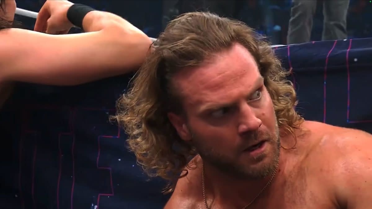 Hangman Adam Page shows a tense expression while reacting to the end of a wrestling event, revealing a mix of emotion and surprise. A blurred figure is partially visible behind him.