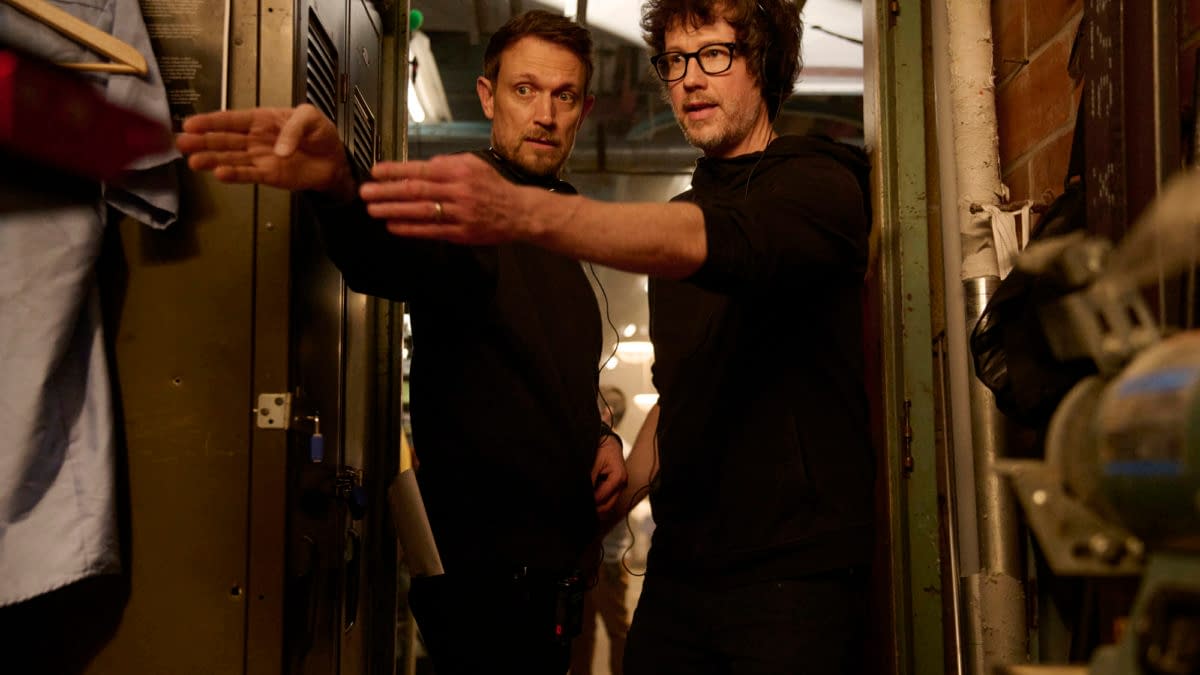 Directors Matt Bettinelli-Olpin and Tyler Gillett discussing a scene on the set of 'Ready Or Not 2: Here I Come', standing in a dimly lit room filled with lockers and equipment.