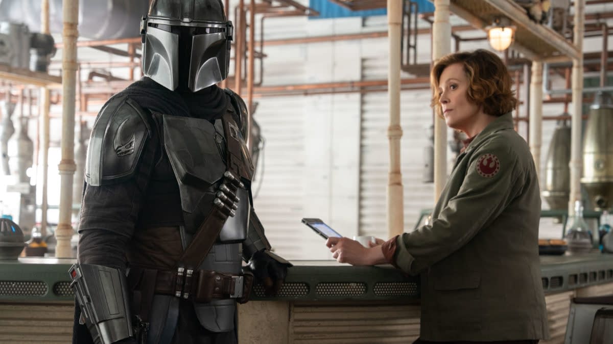 Mandalorian character in full armor stands next to Colonel Ward, who is seated at a bar-like counter, both appearing to engage in conversation. This scene is from Lucasfilm's 'The Mandalorian and Grogu.'