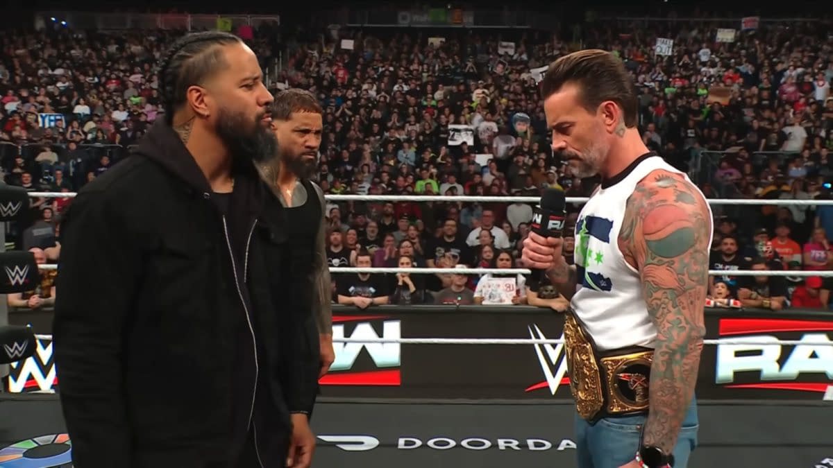The Usos confront CM Punk in the ring during an episode of WWE Raw, with a captivated audience in the background and CM Punk holding a microphone and a championship belt.