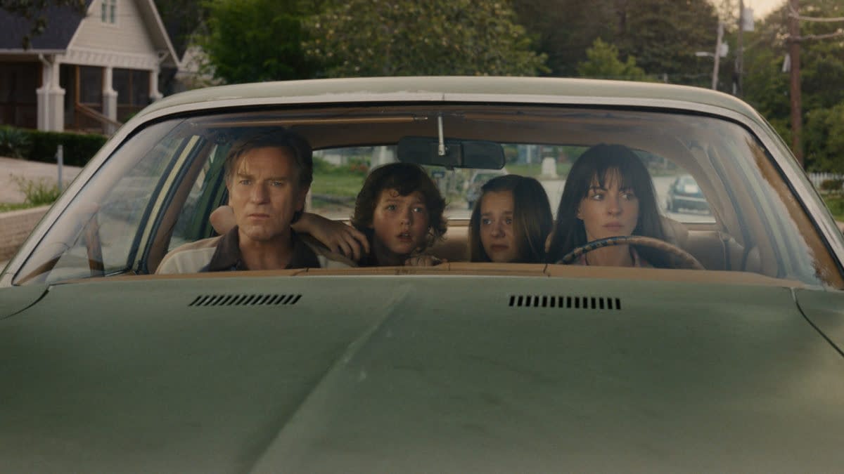 A dramatic scene in a vintage car featuring a man and three children, each displaying varied expressions of concern and intensity while looking out the windshield. The background includes a suburban setting with green trees and houses.