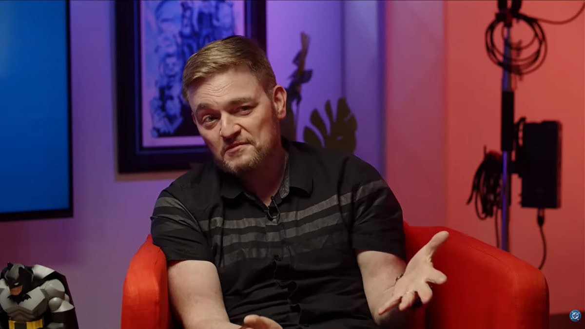 Scott Snyder gives a DC Comics interview. In the background, there are colorful lights and a framed artwork, along with a Batman figurine on a table nearby.