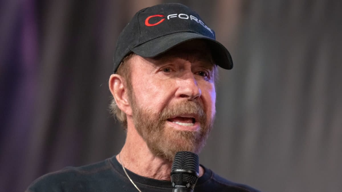 Chuck Norris speaking at the German Comic Con in Dortmund, Germany, on December 1, 2018, wearing a black cap and holding a microphone.