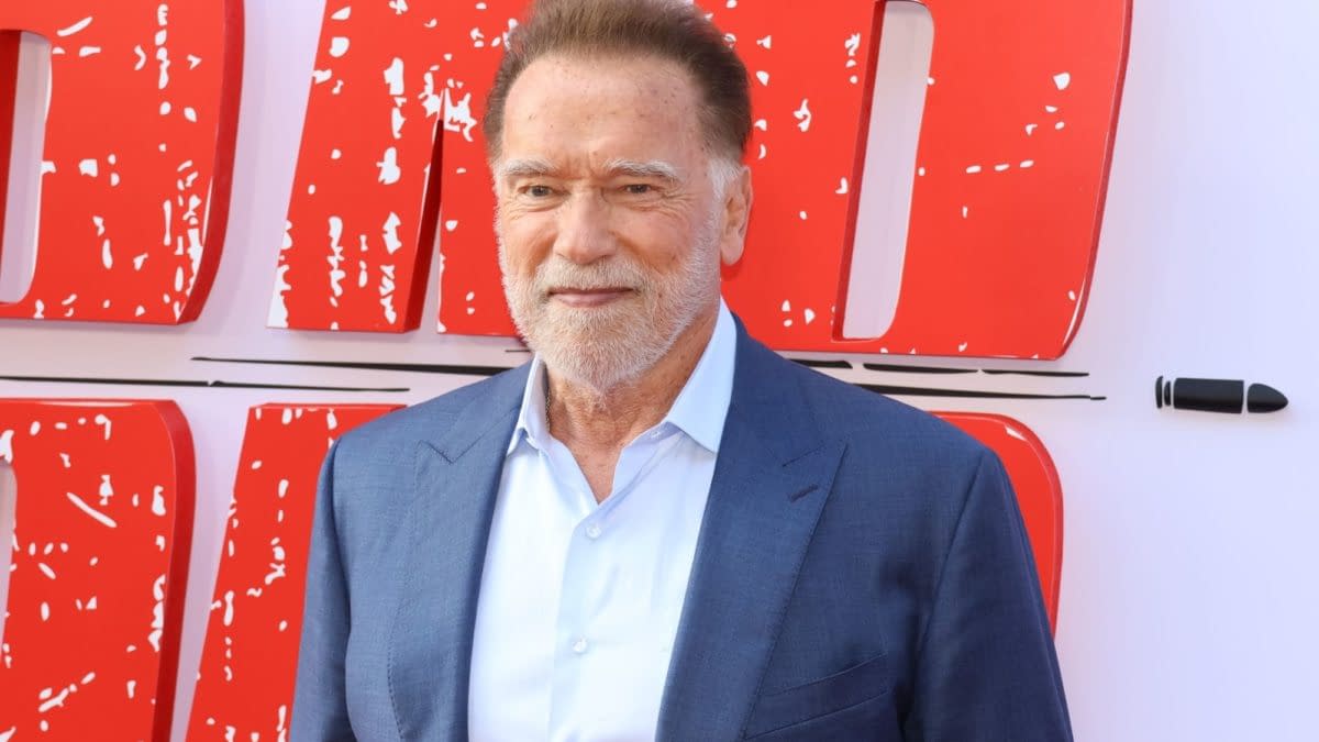 Arnold Schwarzenegger poses at a red carpet event, wearing a blue suit and light shirt, with bold red promotional signage in the background.