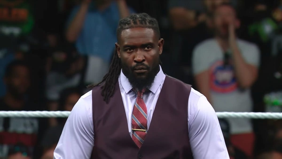 A man with long braided hair wearing a dark vest and light purple shirt stands confidently in a wrestling ring, appearing serious against a cheering crowd in the background. He is dressed for a formal occasion, enhancing his commanding presence.