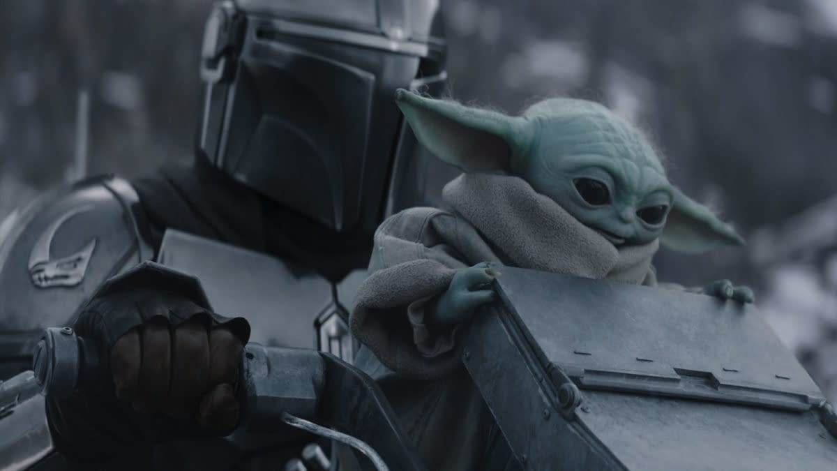 The Mandalorian, wearing a silver helmet and armor, holds onto a vehicle while Grogu, a small green creature with large ears, sits beside him wrapped in a grey robe. The scene conveys a moment of adventure in a snowy landscape.