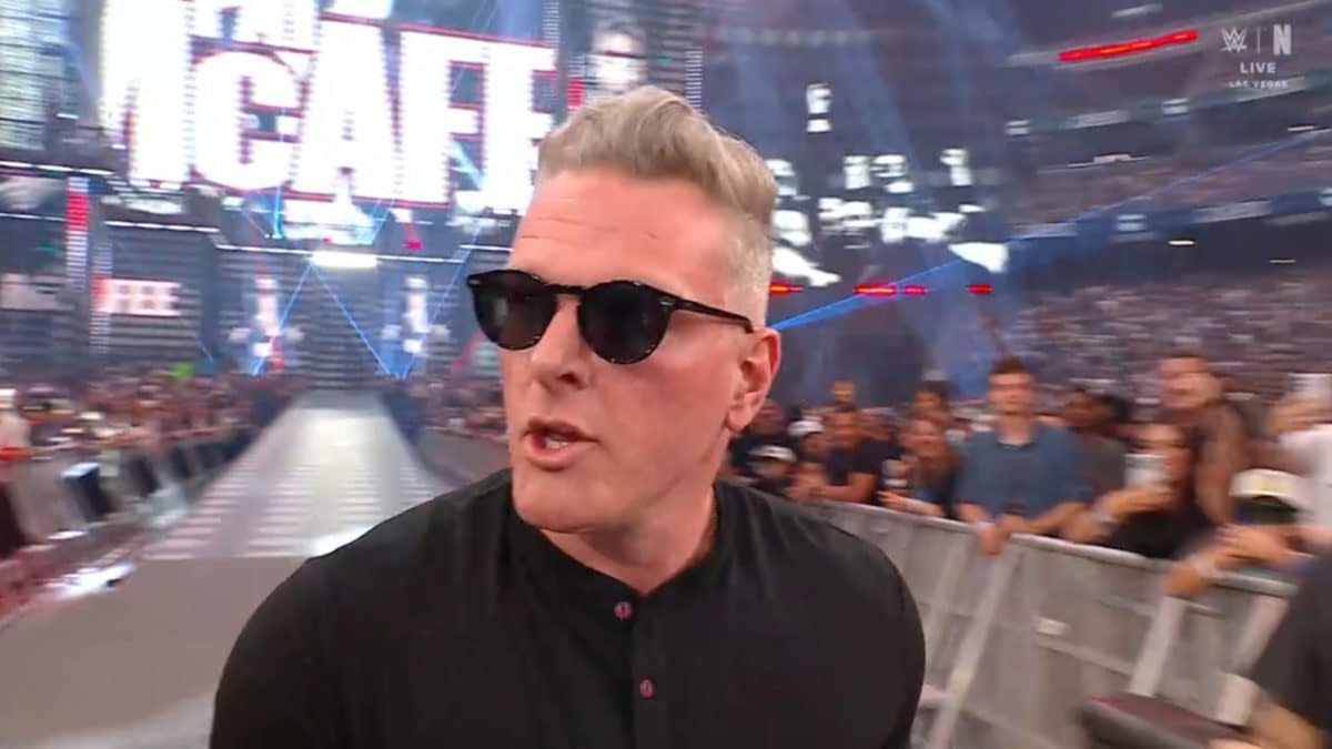 A man with short, silver hair wearing sunglasses walks past a crowd at a wrestling event, with bright lights and a large screen in the background displaying 'The McAfee'.