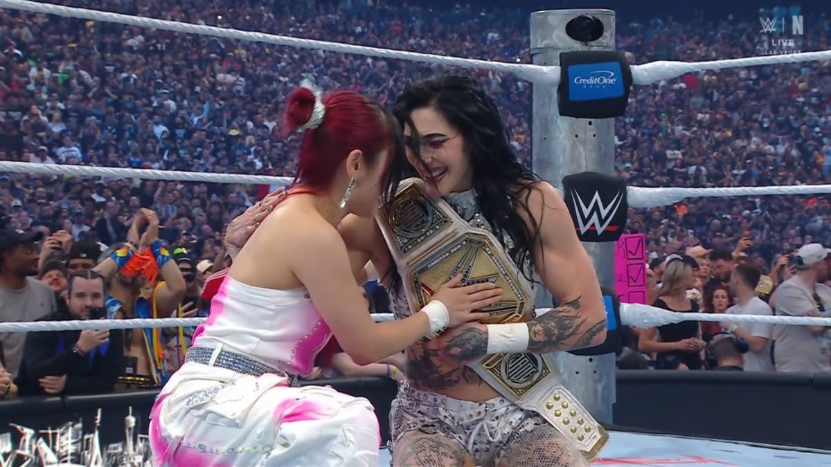 Two women embrace joyfully in a wrestling ring, one holding a championship belt. The crowd is visible in the background, celebrating the moment.