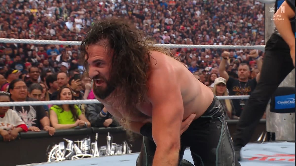 A wrestler with long hair and a beard is shown mid-match, appearing to be in pain as he holds his stomach. The crowd is visible in the background, showing excitement and engagement during the event.