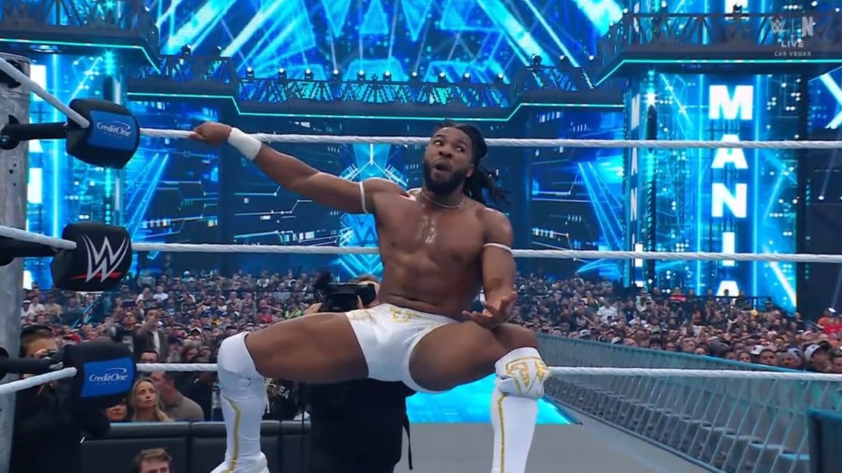 A wrestler celebrating in the ring at WrestleMania, wearing white trunks and striking a dynamic pose against a vibrant blue backdrop filled with lights and an enthusiastic crowd.