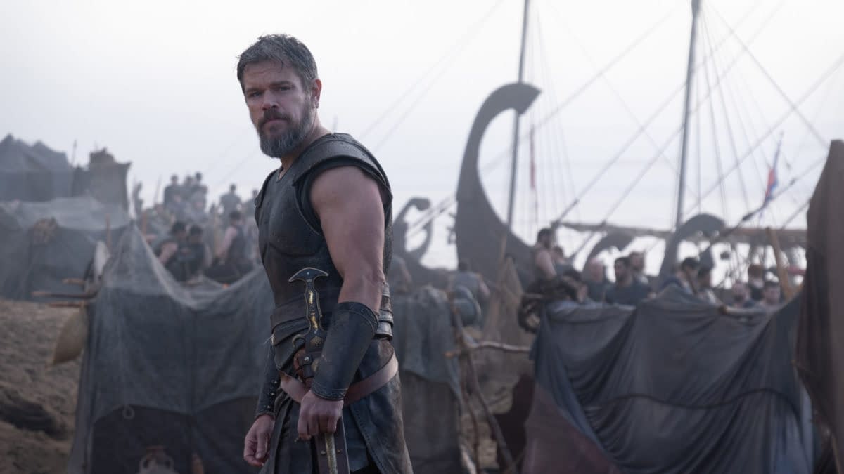 Matt Damon as Odysseus stands in armor at a crowded ancient Greek encampment beside a ship.