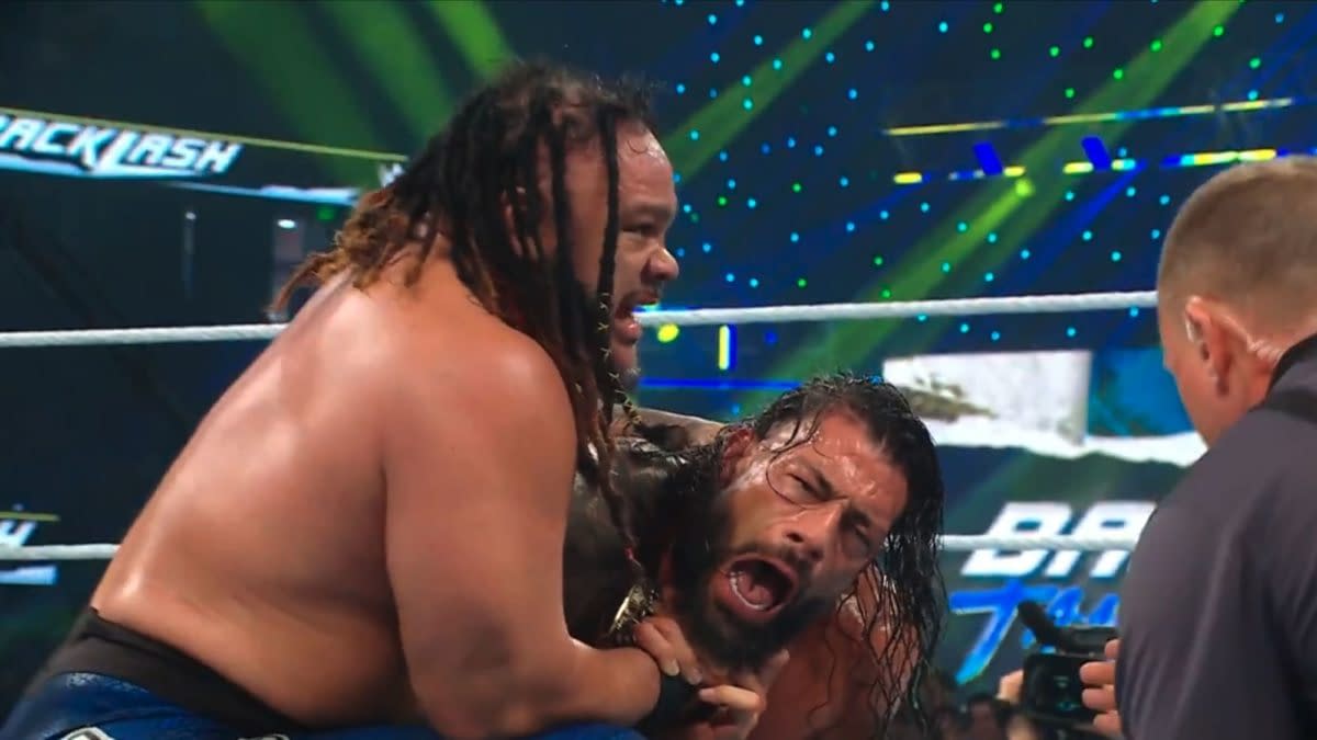 Roman Reigns is trapped in a brutal hold from Jacob Fatu during WWE Backlash. The tense ringside moment shows Reigns grimacing under heavy punishment.