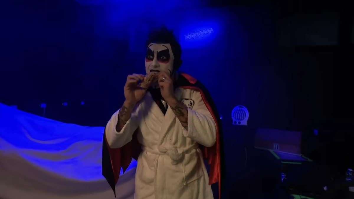 Danhausen, in face paint and a robe, eats a snack under blue lighting on WWE SmackDown.