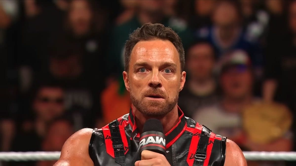 LA Knight stands in the ring on WWE Raw, holding a microphone and looking intensely at the crowd. He wears a black-and-red outfit against a blurred arena audience.