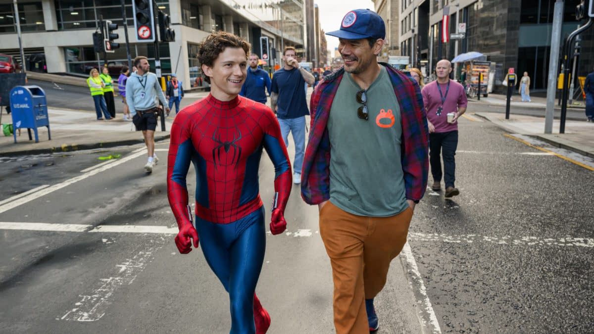 Spider-Man actor Tom Holland walks down a city street with a crew member during filming, smiling in his red-and-blue suit.
