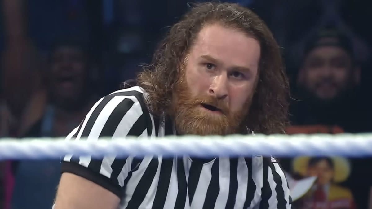 Sami Zayn, wearing a referee shirt, looks intensely toward the action inside the wrestling ring during a WWE title match. The blurred ring rope crosses the foreground.