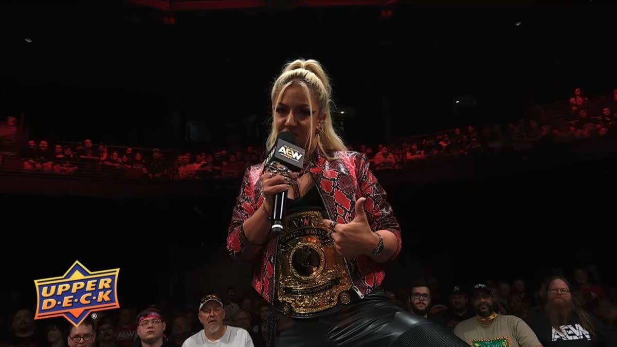 Thekla speaks into an AEW microphone while wearing a championship belt and patterned jacket in a dimly lit arena. She appears to be cutting a promo before Forbidden Door on AEW Collision Summer Blockbuster.