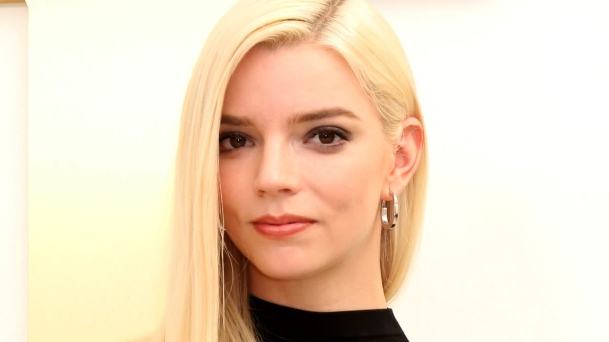 Anya Taylor-Joy poses at an Apple TV press event, wearing a black outfit and silver hoop earrings. Her platinum blonde hair is parted to one side against a bright backdrop.