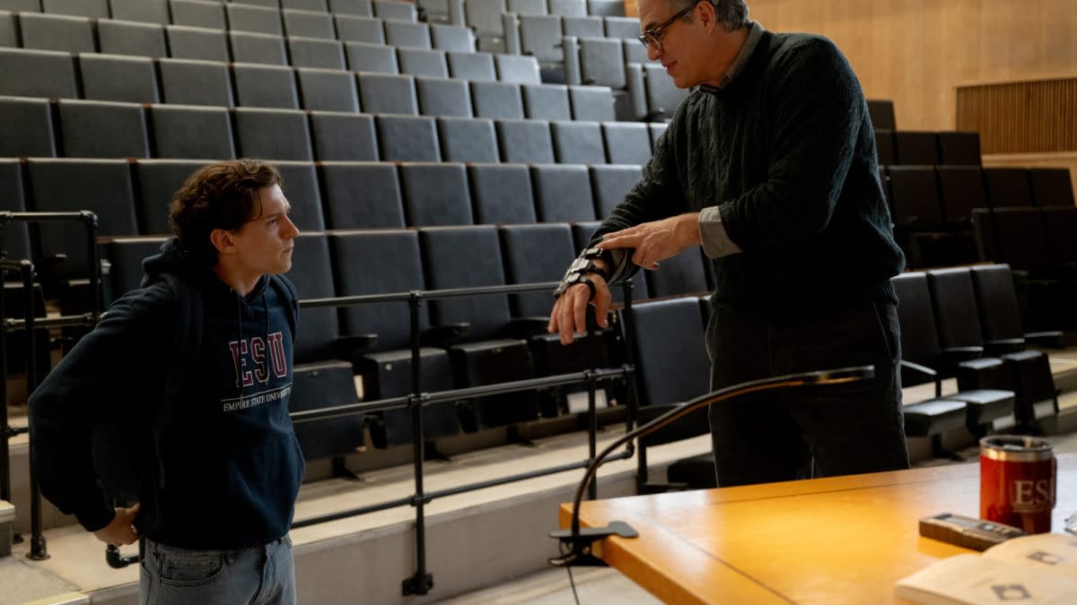 Two men confer in a lecture hall, one showing a wrist device or prosthetic arm to the other. Rows of empty auditorium seats rise behind them.