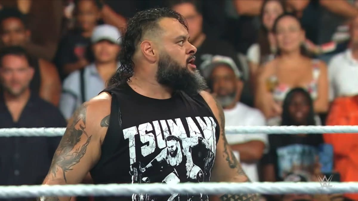 A WWE wrestler with a beard and long wet hair stands in the ring wearing a black “TSUNAMI” shirt, with the crowd blurred behind him.