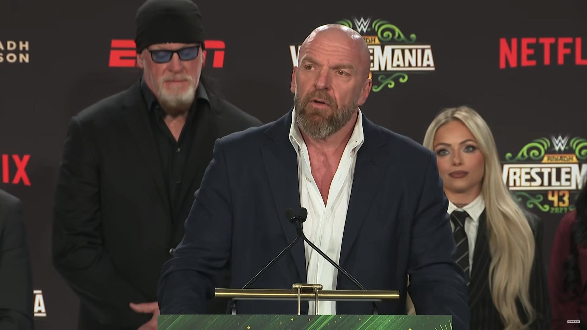 A press conference scene featuring a man in a suit speaking at a podium, with a green WrestleMania 43 logo in the background. Behind him, another man in a black outfit and sunglasses stands, along with a woman with long blonde hair dressed in a suit.