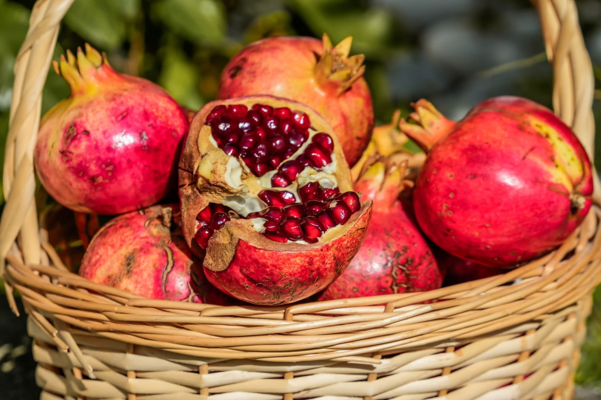 Close-up of ripe pomegranates in a wicker basket, including one split open to reveal glossy red seeds. The rustic still life has a fresh, natural produce feel.