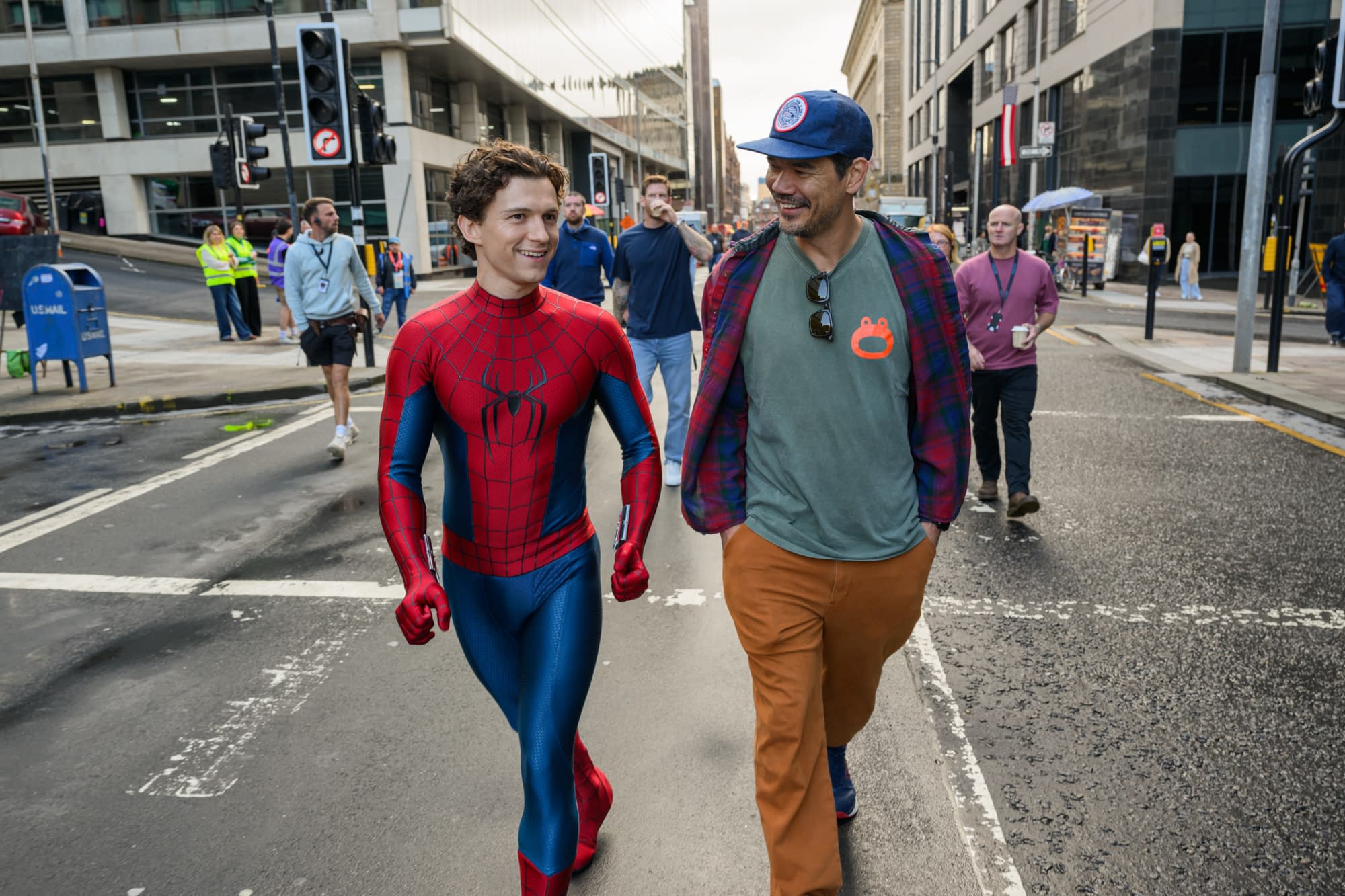 Spider-Man actor Tom Holland walks down a city street with a crew member during filming, smiling in his red-and-blue suit.