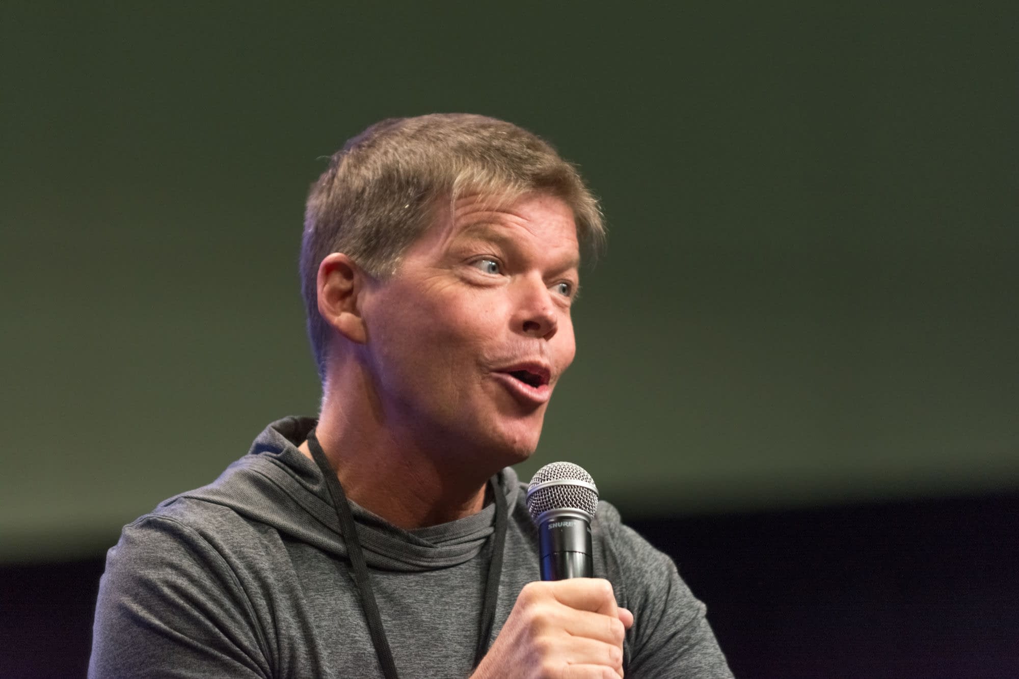 Rob Liefeld speaks into a handheld microphone at a comic convention panel in Los Angeles. He wears a gray hoodie and looks animated mid-sentence.