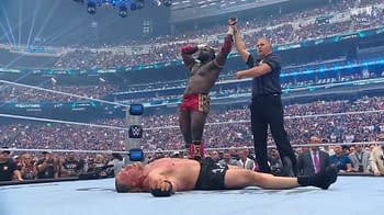 In a wrestling ring, Oba Femi stands triumphantly with his arm raised, having defeated Brock Lesnar, who lies on the mat beneath him. The audience is visible in the background, filled with cheering fans.