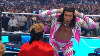 A professional wrestler in a bright pink and white costume makes an exaggerated facial expression and gestures with both hands near his head. The wrestling ring is filled with cheering fans in the background.