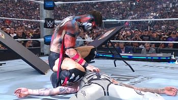 A wrestler with face paint and an intense expression pins another wrestler wearing a silver outfit to the mat during a match in a dramatic arena filled with an excited crowd. The atmosphere is electric with fans in the background cheering and watching intently.